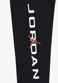 Črni športni rokav z besedo "JORDAN" v debelih belih črkah. Vključuje rdečo silhueto košarkarja. Mehka tekstura tkanine.