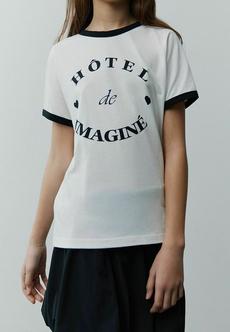 Enfant portant un T-shirt blanc avec des bordures noires et un texte noir disant "HÔTEL de IMAGINÉ" et de petites formes de cœur sur le devant.