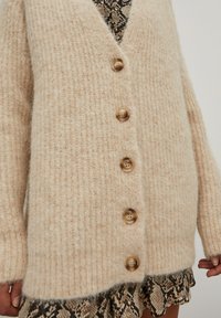 Beige gebreide cardigan met een V-hals, vijf houten knopen, geribbelde textuur en lange mouwen, gedragen over een patroonjurk.