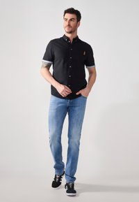 Camisa negra de botones de manga corta con un bolsillo en el pecho, ribete del collar en azul claro; combinada con jeans de mezclilla azules y zapatillas negras.
