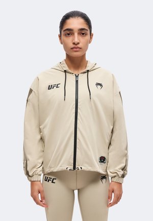 Mujer con sudadera con cremallera beige de la UFC y leggings a juego, de pie mirando al frente con el pelo recogido y expresión neutral.