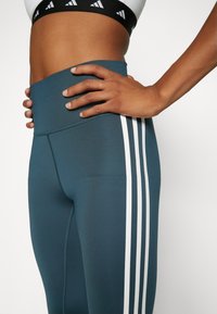 Leggings realizzati in materiale elastico di un verde teal scuro, con una vita alta e tre strisce bianche verticali lungo i lati.
