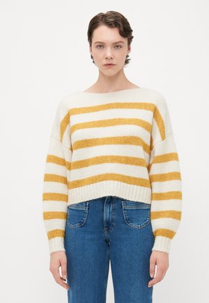 Ung person iført en hvit og sennepsstripet genser med lange ermer og blå jeans, står foran en ensfarget hvit bakgrunn.