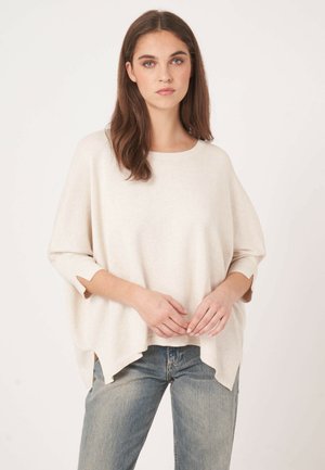 Junge Frau mit langem braunem Haar trägt einen locker sitzenden cremefarbenen Pullover und blaue Jeans, steht vor einem schlichten weißen Hintergrund mit verschränkten Händen.