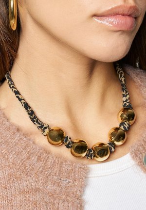 Goldfarbene Perlen, gemischt mit einem schwarz-beigen gemusterten Stoff, bilden eine voluminöse Halskette. Der Stoff ist geflochten und strukturiert.