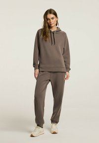 Brązowa bluza z kapturem i dopasowane joggery wykonane z miękkiego materiału. Bluza ma sznurek do regulacji i ściągacze. Joggery są zwężane, w połączeniu z beżowymi trampkami.