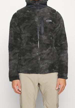 Zwarte, pluizige jas met volledige ritssluiting en borstzak, gedragen door een persoon die beige broek draagt, zichtbaar van taille tot halverwege de dij.