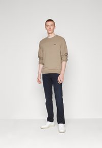 Beige knit sweatshirt with round neckline and embroidered logo, paired with dark blue jeans and white sneakers, standing against a neutral background.