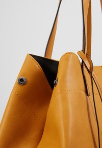 Gros plan sur un sac à main en cuir fauve avec intérieur noir, coutures visibles, boutons-pression métalliques et fines bretelles.