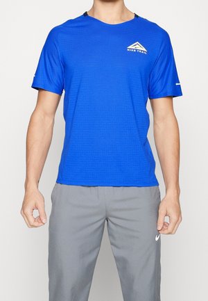 Mann trägt ein blaues Nike Trail Kurzarm-Sportshirt und graue Nike-Hose, steht mit entspannten Armen seitlich am Körper.