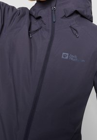 Marinblå vattentät jacka med hög krage, justerbar huva och frontdragkedja. Har en logotyp på vänster bröst och snörjusteringar.