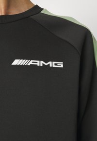 Czarna i zielona bluza z białym logo AMG nadrukowanym na lewej górnej części klatki piersiowej, zbliżenie na ramię i dekolt.