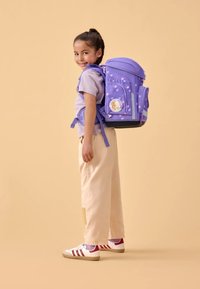 Mochila morada con puntos, correas ajustables, bolsillos laterales y una insignia de personaje. La lleva puesta un niño con un atuendo ligero y zapatillas deportivas.