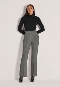 Frau steht mit hinter dem Rücken verschränkten Händen, trägt einen schwarzen Rollkragenpullover, gemusterte hoch taillierte Schlaghosen und schwarze Stiefeletten mit Absatz.