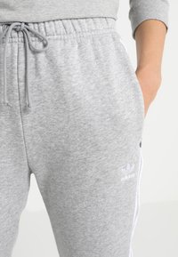 Pantalons de survêtement en coton gris avec une taille élastique, un cordon blanc et des rayures blanches sur les côtés. Comprend le logo Adidas sur la cuisse.