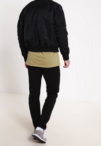 Giacca bomber nera, camicia verde oliva, pantaloni neri e sneaker grigie. Caratteristiche includono polsini e vita a coste sulla giacca.