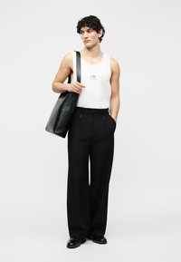 Άνδρας που φορά λευκό αμάνικο τοπ, μαύρο παντελόνι με φαρδιά μπατζάκια, μαύρα παπούτσια, κρατώντας μεγάλη μαύρη τσάντα ώμου, στέκεται μπροστά σε απλό φόντο.