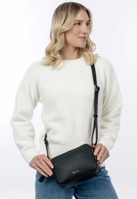 Borsa a tracolla nera texture a forma rettangolare, con tracolla regolabile e dettagli del logo discreti. Indossata con un maglione bianco in maglia e jeans.