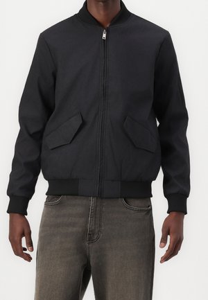 Mann trägt eine schwarze Bomberjacke mit Reißverschluss, gerippten Bündchen und zwei schrägen vorderen Klappentaschen, kombiniert mit ausgewaschenen schwarzen Jeans.