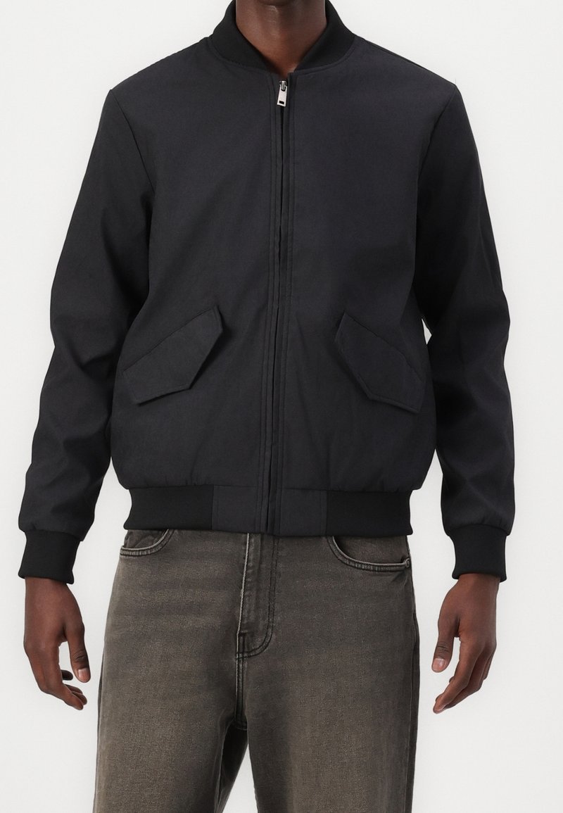 Homme portant une veste bomber noire zippée avec des poignets côtelés et deux poches avant inclinées à rabat, associée à un jean noir délavé.
