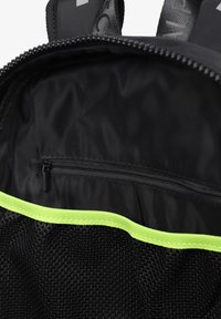 Interior de una mochila de malla negra con un ribete verde neón, un bolsillo con cierre y un forro de tela suave. Se pueden ver las correas ajustables.