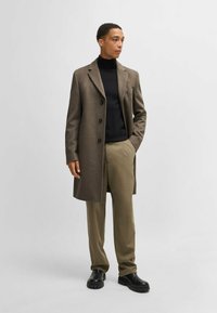 Cappotto di lana marrone con rever a scollo e quattro bottoni neri, indossato sopra un dolcevita nero e pantaloni khaki, abbinato a scarpe nere.