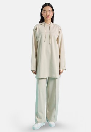 Donna in piedi con una felpa beige ampia con coulisse, pantaloni a gamba larga abbinati e sneakers bianche su uno sfondo chiaro e uniforme.