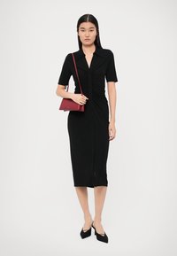 Robe noire ajustée à manches courtes, avec un boutonnage à l'avant, un détail plissé sur le côté, longueur genou. Associée à un petit sac bandoulière rouge et des talons noirs.