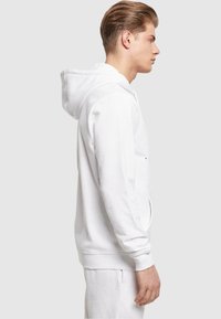 Hvid bomulds hoodie med kengurulomme, snøre i hætten og ribbestrikkede håndled. Glat tekstur og afslappet pasform, vist fra siden.