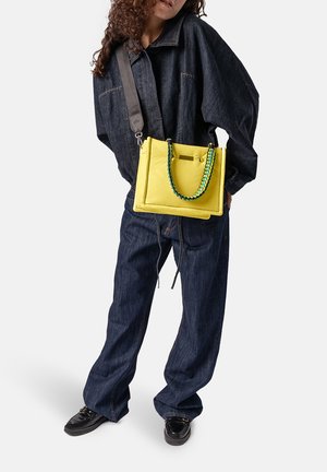 Person trägt dunkelblauen Jeans-Overall und schwarze Schuhe, trägt eine leuchtend gelbe Handtasche mit geflochtenem Henkel über der Schulter.