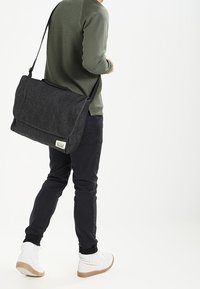 Bolso mensajero de denim negro con tapa, correa ajustable y etiqueta. Lleva puesto un suéter texturizado verde y pantalones negros. Zapatillas blancas.