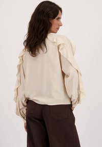 Femme aux cheveux ondulés foncés portant un chemisier crème avec des manches volantes et un pantalon taille haute sombre, tournée de côté devant un fond uni.