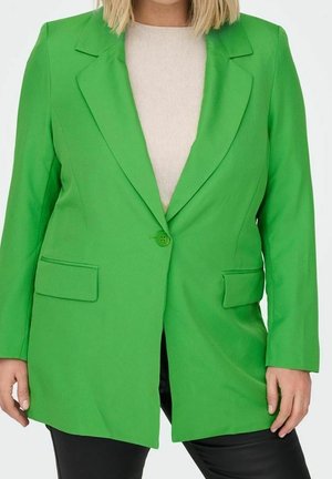 Kobieta ubrana w jaskrawozieloną marynarkę na jedno zapięcie, na białą bluzkę i czarne spodnie, stojąca z rękami swobodnie opuszczonymi wzdłuż ciała.