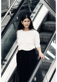 Une personne portant un pull en tricot blanc et une jupe noire se tient sur un escalator métallique, avec en arrière-plan une texture d'escaliers et de rampes en verre.