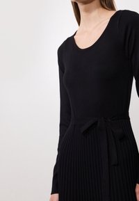 Robe noire en maille avec un décolleté rond, des manches longues et une jupe côtelée. Elle présente une ceinture nouée pour un ajustement réglable.