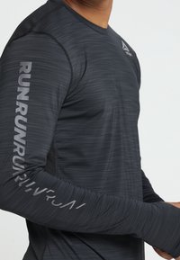 Zbliżenie na osobę w ciemnoszarej, długiej sportowej koszulce z napisami "RUN" powtarzającymi się na rękawie oraz małym logo na klatce piersiowej.