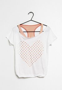 Bílý krátký rukáv T-shirt s výřezem ve tvaru srdce, který je vytvořen z otvorů. Má měkkou texturu a pod ním jsou viditelné vrstvené růžové popruhy.