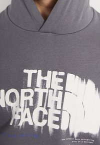 Sudadera gris con un cuello alto, diseño oversize y una impresión gráfica blanca que incluye "THE NORTH FACE" y detalles de texto adicionales.