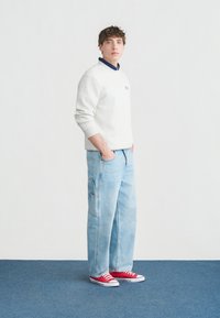 Νέος άνδρας που στέκεται με τα χέρια στις τσέπες, φοράει λευκό φούτερ, ανοιχτό μπλε τζιν παντελόνι με φαρδύ μπατζάκι, κόκκινα αθλητικά παπούτσια και μπλε μπλούζα με γιακά από κάτω.