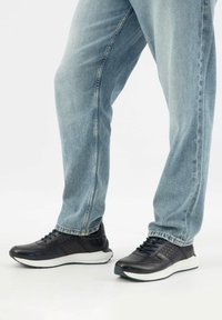 Osoba oblečená v modrých dž ínách a černých teniskách s bílou podrážkou, stojící na jednoduchém světlém pozadí.