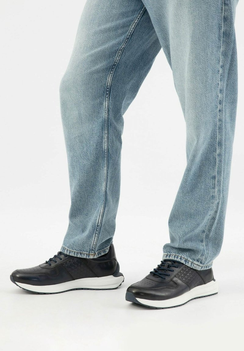 Osoba oblečená v modrých džínách a černých teniskách s bílou podrážkou, stojící na jednoduchém světlém pozadí.