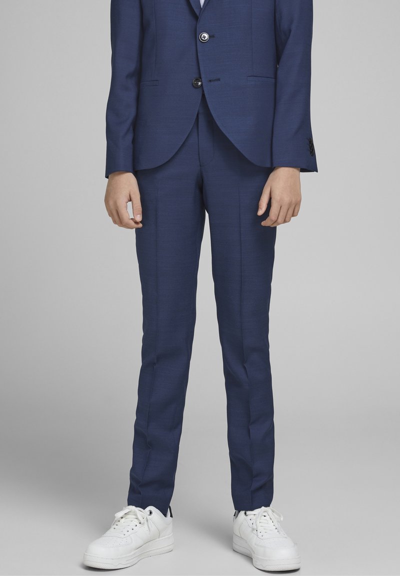 Personne portant un costume bleu marine ajusté avec une veste à deux boutons et des baskets blanches, se tenant devant un fond gris uni.