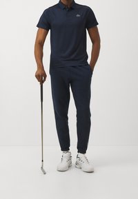 Mężczyzna w granatowej koszulce polo i dresowych spodniach, trzymający kij golfowy w jednej ręce, stoi na tle białego tła.