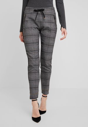 Woman wearing gray plaid drawstring pants with zippered pockets and black high-heeled ankle-strap shoes, standing on a light background.
