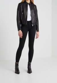 Nainen, jolla on päällään musta nahkatakki, valkoinen paita, mustat kapeat farkut ja mustat nilkkasaappaat, seisoo yksinkertaista taustaa vasten.