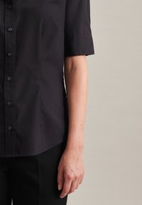 Camicetta nera con bottoni, maniche corte, texture liscia, orlo leggermente arrotondato e un logo sottile sulla parte anteriore. Abbinata a pantaloni scuri.