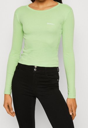 Person trägt ein eng anliegendes hellgrünes Langarmshirt mit kleinem weißem Schriftzug und hoch taillierte schwarze Hose mit Doppelknopfverschluss.