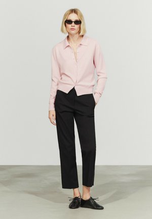 Femme portant des lunettes de soleil noires, un cardigan rose clair boutonné, un pantalon noir court et des chaussures noires à lacets, debout avec une main dans la poche.