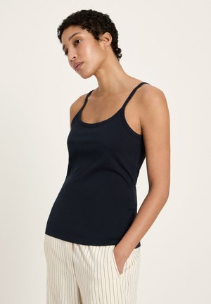 Woman with short curly hair wearing a navy blue tank top and white pants with black vertical stripes, standing with one hand in pocket.