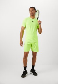 Conjunto deportivo de camiseta y pantalones cortos en verde neón con acentos negros, sosteniendo una raqueta de tenis. La tela parece suave y elástica.
